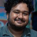 Close-up portrait of a smiling man with curly hair wearing a casual checkered shirt indoors.