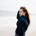 Young woman in a stylish outfit enjoying a breezy day at the beach, with ocean waves in the background.